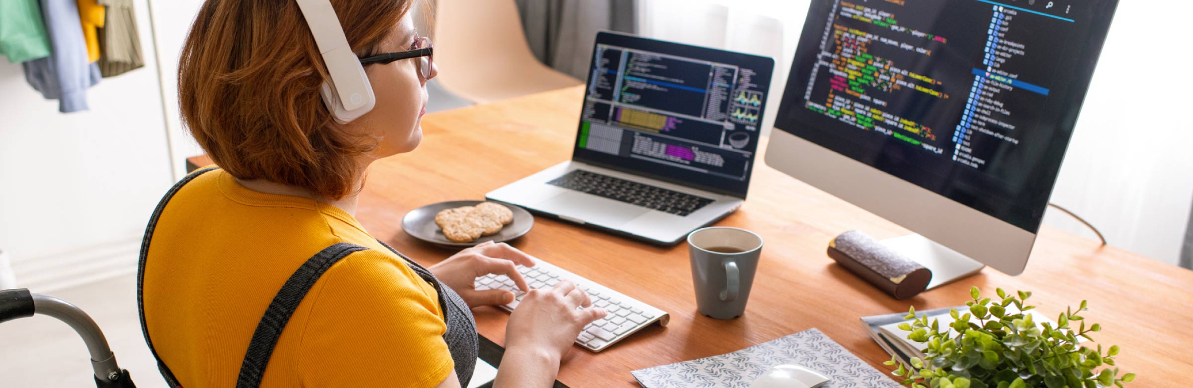 A woman wearing headphones sits at a desk in front of a laptop and computer monitor testing software applications.